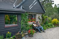 Couple sitting on a rustic wooden carriage wheel seat in front of house surrounded with planting