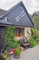 House front with old skis attached. Planting around house includes 
window boxes and oak half barrel with red Pelargoniums, plus bed under window and rustic wooden carriage wheel
 seat

