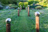 View up grass path in middle of avenue of stainless steel mirror globes
 mounted on wooden posts. Main plant in meadow is Camassia subsp. leichtlinii
