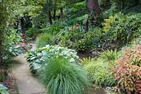 Pathway passes woodland border and rocky pool, with hostas, ferns and ornamental grasses. 
