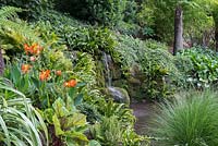 A cascade of water splashes into a pool edged in hart's-tongue ferns, Bergenia, Hosta, ornamental grasses, ivy and Canna.