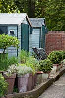 Potted herbs stand outside painted wooden sheds.