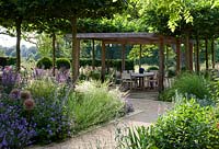 Wooden pergola creates shaded dining area, surrounded by trees, flowering perennials and ornamental grasses. 