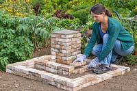 Woman adding bricks to create two L-shaped tiered beds around the corner pedestal. 