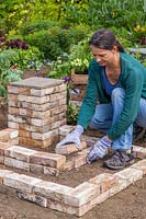 Woman adding bricks to create two L-shaped tiered beds around the corner pedestal.