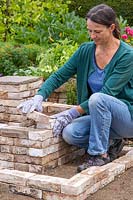 Woman adding bricks to create two L-shaped tiered beds around the corner pedestal.