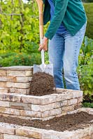 Woman adding well-draining soil to tiered brick garden using a spade. 