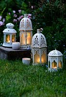 An eclectic mix of white candle lanterns in garden at dusk. 