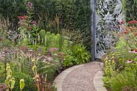 A curved gravel path with stone edges leading through borders to an ornate metal gate with perennial planting including Helianthus salicifolius, Phlomis russeliana and Monarda 'Jacob Cline' in the 'Don't Chop Me Down' Garden at Tatton RHS Flower Show 2018