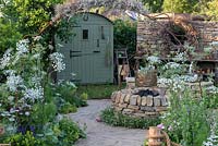 A romantic country garden planted with mostly medicinal plants, a shepherd's hut, an eclectic collection of objects, and a dry-stone firepit, set into a circular path of clay setts. The Naturecraft Garden, designed by Pollyanna Wilkinson, RHS Hampton Court Palace Garden Festival, 2019. 