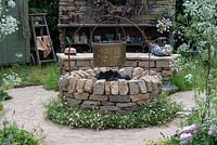 A romantic country garden with a dry-stone firepit, set into a circular path of clay setts, and separated by a collar of fleabane. The Naturecraft Garden, designed by Pollyanna Wilkinson, RHS Hampton Court Palace Garden Festival, 2019.