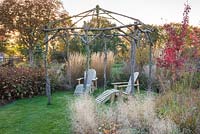 Two wooden recliners under rustic arbour in wildlife friendly garden with autumnal colour.

