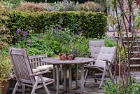 A sheltered seating area, tucked away below a yew hedged and raised beds studded with purple allium and hardy geraniums.