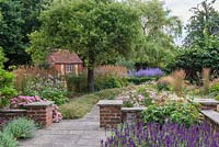 From stone terrace, path leads to parterre with perovskia,  passing apple tree above banks of erigeron, Japanese anemones and feather reed grass on the left. To the right, border of achilleas, grasses and coneflowers.