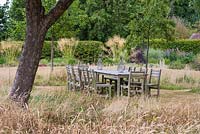 Table and chairs set in shade of apple trees, surrounded in uncut grass. Beyond borders studded with large clumps of Stipa gigantea.