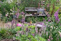 Gardening tools and cushion on rustic garden bench nestled amongst perennial planting along winding gravel path. RHS Hampton Court Festival 2019. 