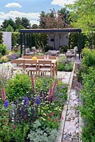 Water rill with pebbles running along perennial borders. Timber and steel open garden room with Green living roof and hardwood table and chairs in contemporary garden. RHS Hampton Court Festival 2019.