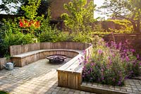 Seating area with fire pit and round wooden bench surrounded by Erysimum 'Bowles's Mauve', Prunus avium 'Stella' and Cornus alba 'Sibirica' 