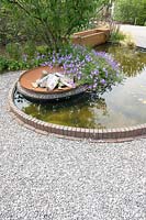Round pond edged with bricks set in gravel. Within the pond a fire pit with Geranium 'Rozanne'