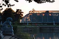 View from terrace with lion statue across pond towards Palm House at dusk
