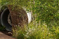 Naturalistic planting with Leucanthemum vulgare - Ox-eye daisies and Betula pendula - Silver Birch - and a woven willow arch covering a large water pipe. RHS Hampton Court Palace Garden Festival 2019. Sponsor: Thames Water.