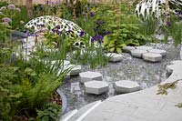 View of sculptures and cottage garden planting, with hexagonal stepping stones over the pond, bordered by centranthus ruber 'Albus', rodgersia, and iris sibirica. Garden: The Manchester Garden. Chelsea Flower Show 2019.