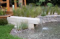 A concrete water feature and pond made from dry stone walling bordered by a gravel path with Stipa tenuissima bordering the pond - The Harmonious Garden of Life Garden - Sponsor: Mr Robert and Mrs Sue Cawthorn Margheriti Piante, Italy
