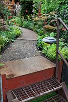 View of path made of rusty metal and porous spoil from the quarry, with metal steps, bordered by geum 'Totally Tangerine' and mixed grasses. Garden: Walker's Forgotten Quarry. Sponsor: Walkers Nurseries. Chelsea flower show 2019.