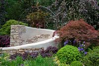 Fagus sylvatica 'Purple Mountain' trees providing the backdrop to a dry stone wall seating area with decorative sundial dome, plants include Loropetalum, Acer palmatum, Carex and Buxus sempervirons topiary - Miles Stone: The Kingston Maurward Garden, RHS Chelsea Flower Show 2019.