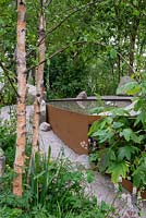 Gravel path leading round a raised corten-steel pool in a woodland garden with Betula nigra - Family Monsters Garden, RHS Chelsea Flower Show 2019