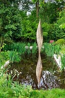 The Savills and David Harber Garden. Calm green  planting centred around rectangular pond with bronze-coloured metal ribbed sculpture shard which is reflected in the water. Planting includes: Anthriscus sylvestris - Cow Parsley, Iris pseudacorus and Ranunculus acris - Meadow Buttercup. Sponsor: Savills and David Harber
