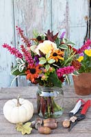 Posy or Tussie Mussie with a colourful mix of flowers and foliage in a jam jar. On a wooden table with nuts, Winter Squash and secateurs
