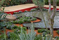 View over the Essence of Australia Garden, shows curved wooden decking and curved reflective water feature pond. Australian outback planting: Brachyscome amongst red-brown earth. Sponsor: Tourism Victoria, Tourism Northern Territory