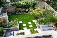 Overview of garden from second floor, looking over lawn with white stone paving and patio, with seating areas. Raised beds filled with mixed plantings. Garden surrounded by brick walls topped with trellis.
