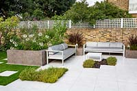 View across patio with white paving, with planting pockets for Chamaemelum nobile - Chamomile and Thymus serphyllum 'Russetings' - Thyme, towards outdoor sofas and raised bed full of plants such as Agapanthus 'Queen Mum'