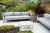 Seating area with grey garden sofas, privacy from trellis top to brick walls. Phormium 'Amazing Red' and Stipa tenuissima in grey raised bed and Thymus serphyllum Russetings - Thyme - and Ajuga reptans 'Atropurpurea' in foreground