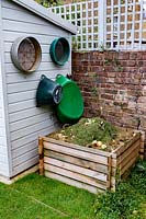 Small wooden compost heap against boundary wall with trellis top. Sieves and plastic trugs hanging from side of wooden shed