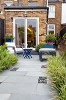 Small contemporary garden in West London, looking towards kitchen dinning room. Planting includes Carex Ice Dance, Heuchera Obsidian, with garden sofa and stool in background.