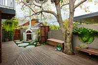 A wide view of a backyard, with a  cement rendered pizza oven in the back corner part hidden a slat hardwood timber screen and groups of pots and a Jacaranda tree growing through the deck.
