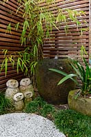 Detail of a family group of hand carved sculptures in front of a timber slat screen with a carved stone pot water bowl, set in amongst a planting of mini Mondo grass next to an in situ cast concrete stepping stone.