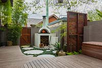 A wide view of a backyard, with a bespoke cement rendered pizza oven in the back corner of a backyard showing a slat hardwood timber screen, a timber deck, a clump of bamboo, groups of pots and a Jacaranda tree growing through the deck.