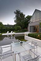 View across dining area to recliners on paved patio surrounded by pool in modern garden. 