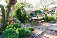 Picnic bench on gravel surface separated by railway sleepers. Seating area surrounded by fruit trees with Rosa - Climbing Rose - and woven spheres, clipped Buxus - Box - topiary balls, self-seeded Erigeron karvinskianus and Geranium palmatum  
