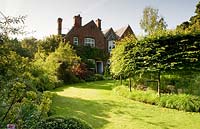 View over lawn to the house, with circle of pleached Carpinus betulus - Hornbeam - to right and curved flower bed to the left

