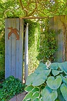 Close board fencing with rabbit wall ornament, next to open, wooden gate by flowering Hosta 