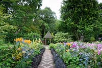Ophiopogon planiscapus 'Nigrescens' edges the path running through the Canal Garden, leading toward the folly framing a white drinking fountain in the Dell Garden at York Gate garden in July. Beds are planted with phlox, orange lilies, Verbena bonariensis and eryngiums.