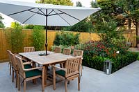 Stone patio area in West London garden, with wooden table, chairs and parasol. In the background there is a border featuring Monarda Fire Ball, Acer palmatum, Helenium Moerheim Beauty, bamboo in terracotta containers, Euonymus Jean Hughes.
