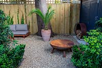 Lower gravel covered patio area in West London garden, with sofas and fire pit - planting includes Euphorbia wallichii, Phoenix palm in terracotta pot.