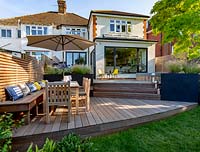 Contemporary garden in Wimbledon with decking on two levels. The lower section includes a bespoke built in bench with cushions, a table with parasol and chairs. In the background are grey raised beds with perennials.