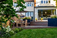 Contemporary garden in Wimbledon with decking on two levels. The lower section includes a bespoke  built in bench with cushions, a table with parasol and chairs alongside grey raised beds with perennials.